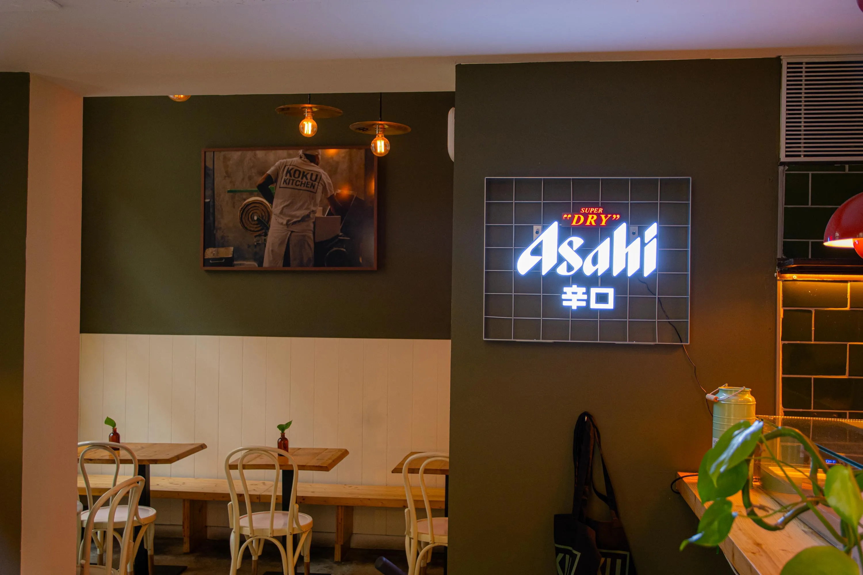 An Asahi neon sign and a framed photo of noodles being made on the green walls of Koku Kitchen Gràcia