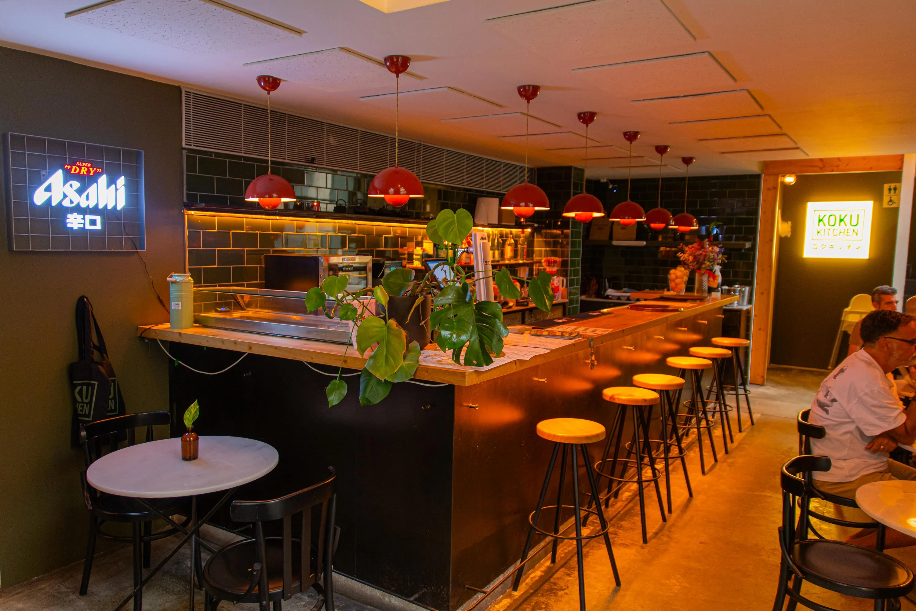 The wooden bar of Koku Kitchen Gràcia under a row of red lamps, with an Asahi neon sign on the wall
