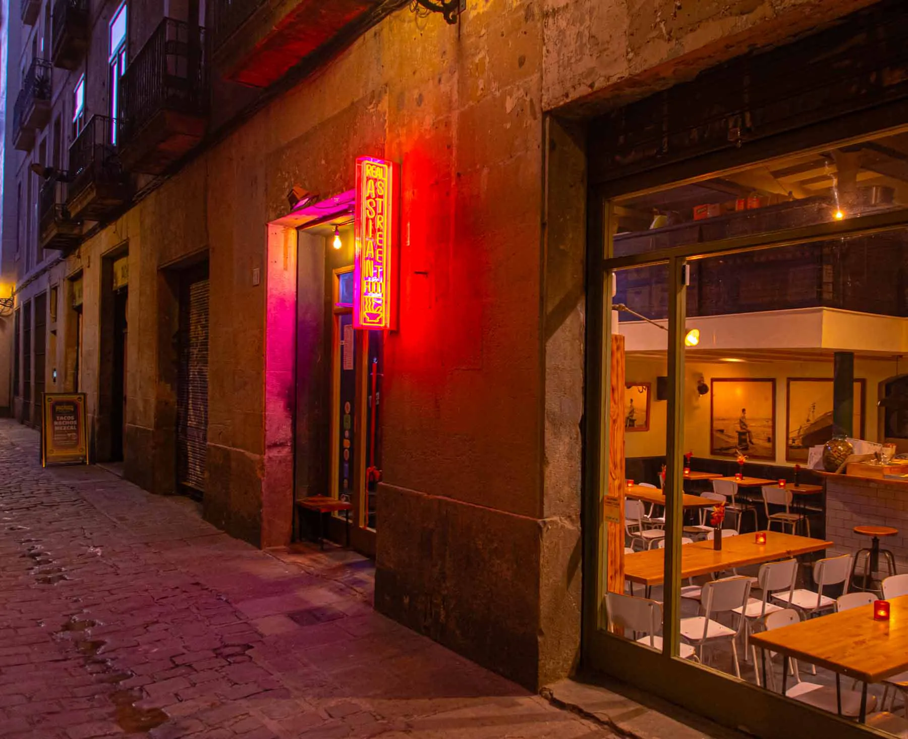 El carrer davant de Koku Kitchen El Gòtic, amb el neó vermell i el menjador a través del vidre